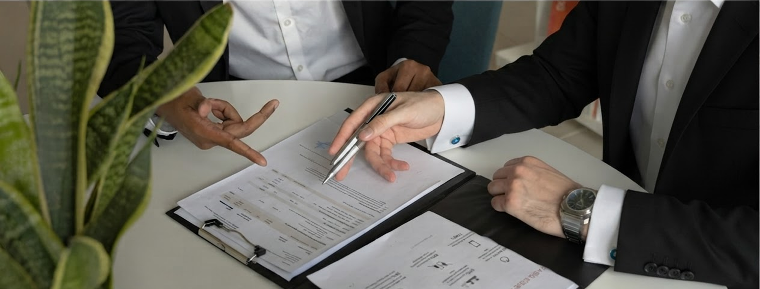 Two professionals in suits sign a document on a clipboard at a round table, one hand offering a pen while a plant sits in the foreground.