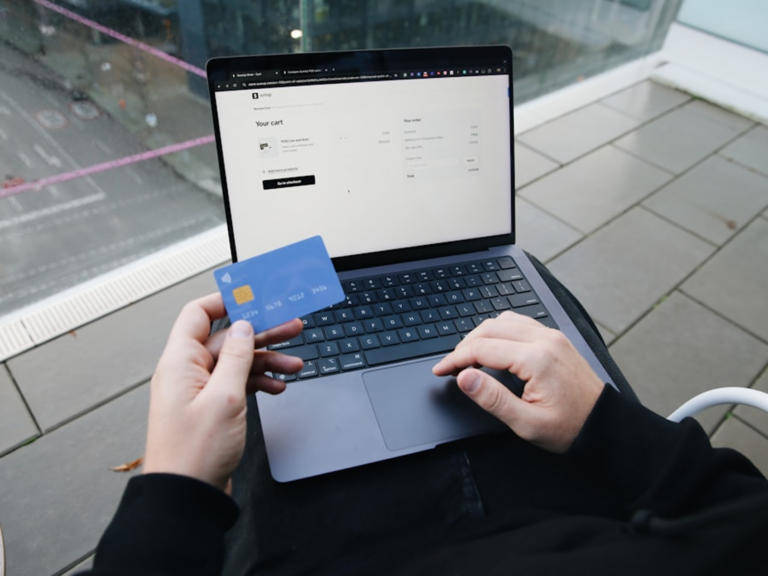 Person outdoors using a laptop while holding a blue credit card near the keyboard, screen shows a checkout page.