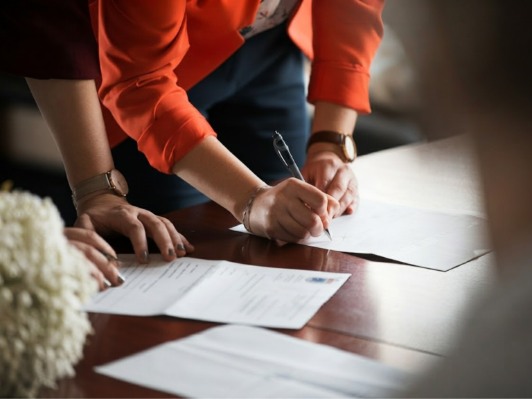 ImageWithFallback - Webtonic - Webtonic Hands with pens signing documents on a wooden table, orange-sleeved arms visible in the background