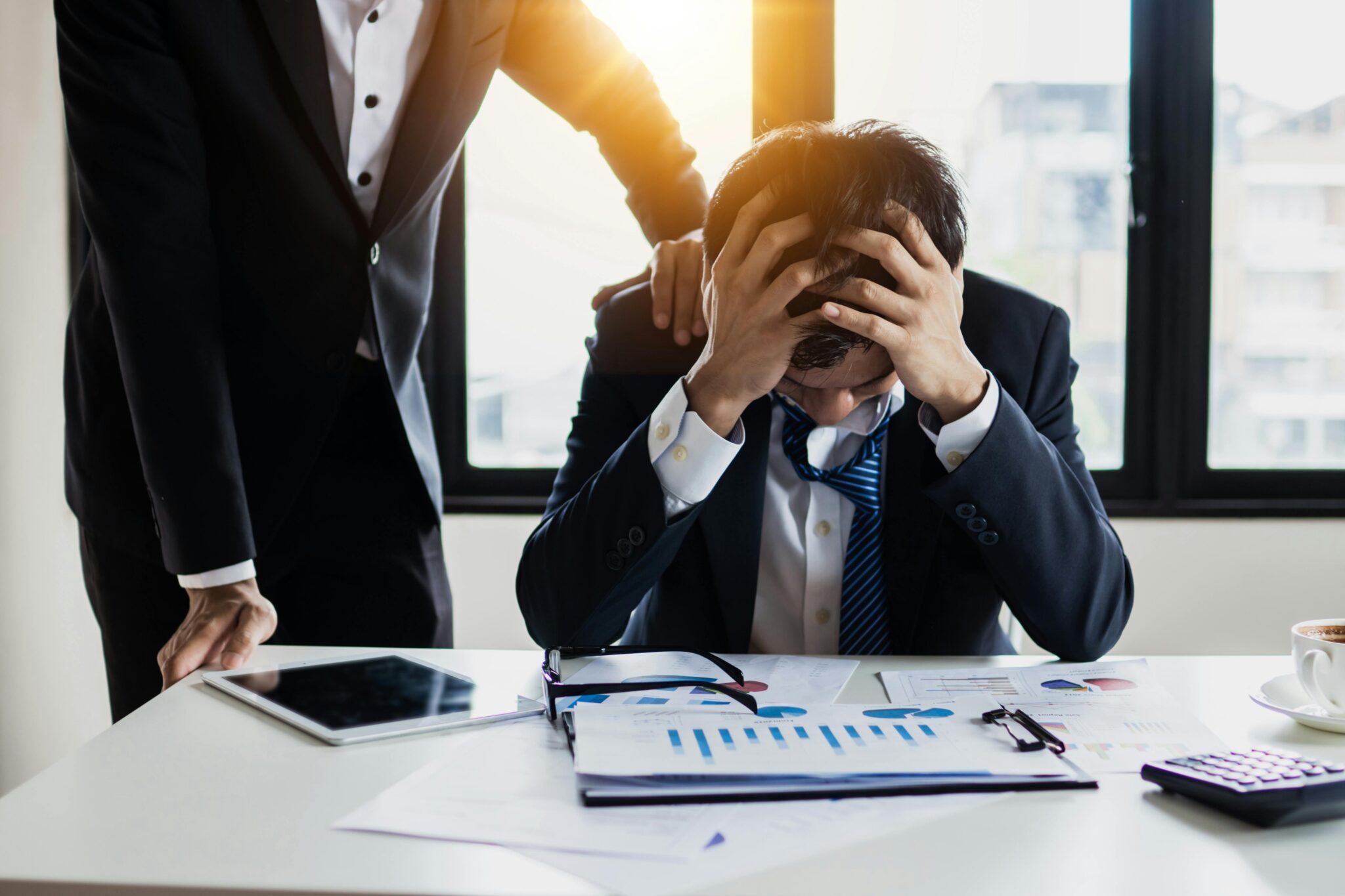 pexels-khwanchai-4175021-1-2048x1365 - Webtonic Man in a suit sits at a desk, holding his head in frustration amid scattered charts and documents while another person stands nearby.