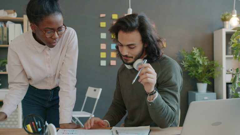 vitaly-gariev-juTM2daTxN0-unsplash - Webtonic - Webtonic Two colleagues collaborate at a desk, reviewing charts and notes with a laptop nearby and colorful sticky notes on a dark wall behind them.